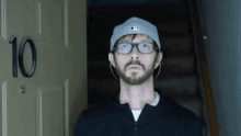 a man with a beard wearing glasses and a hat is standing in front of a door .