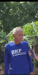 a man in a blue shirt with the word bkp on it is standing in front of a marijuana plant .