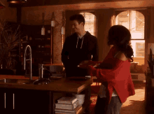 a man and a woman are standing in a kitchen talking to each other . the woman is wearing a red sweater .