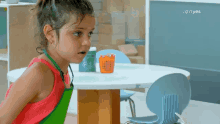 a little girl wearing a green apron is standing in front of a table with a basket of pencils on it .