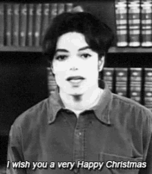 a black and white photo of a man saying `` i wish you a very happy christmas '' in front of a bookshelf .