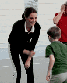 a woman in a black sweater and white pants smiles while talking to a young boy in a green shirt