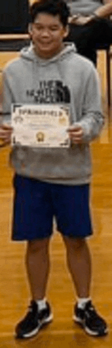 a young man in a gray hoodie and blue shorts is holding a certificate on a wooden floor .