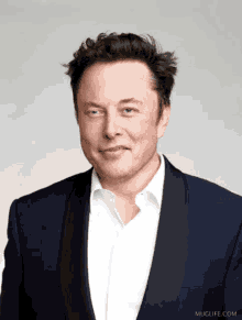 elon musk is wearing a blue suit and white shirt and muglife.com is behind him