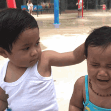 a little girl in a blue tank top is crying while a little boy in a white tank top holds her head