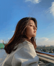 a woman in a grey sweatshirt stands on a balcony with a blue sky in the background