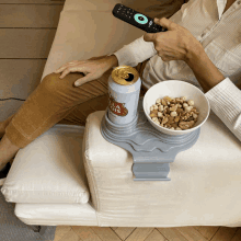 a person sitting on a couch holding a remote control and a can of stella artois beer