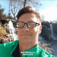 a man wearing glasses and a green jacket stands in front of a waterfall with the words waterfalls are love above him