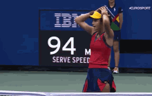 a woman wearing a red tank top and a yellow hat is standing on a tennis court with her hands in the air .