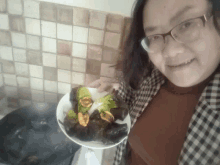 a woman wearing glasses holds a plate of food in front of a stove