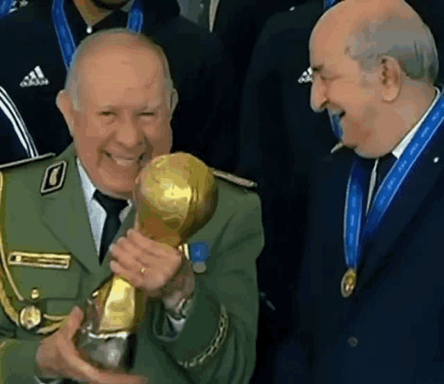 a man in a military uniform is holding a trophy and smiling while another man looks on .