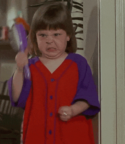 a little girl in a red and purple shirt is holding a brush and making a funny face .