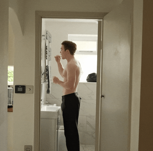 a shirtless man brushing his teeth in a bathroom next to a sink