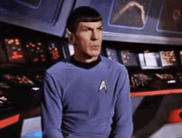 a man in a blue shirt with a star trek logo on his chest is sitting in front of a control panel .