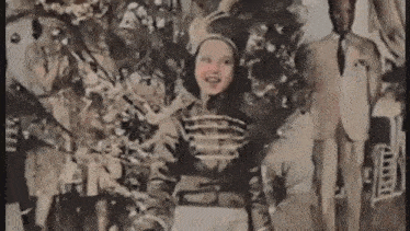 a little girl in a parade uniform is standing in front of a christmas tree with a man in a suit behind her .