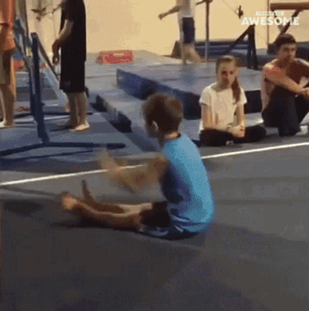 a boy in a blue shirt is sitting on the floor in a gym with a girl sitting on the floor watching him