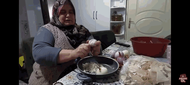 a woman in a hijab is preparing food in a frying pan on a table next to a bag of noodles