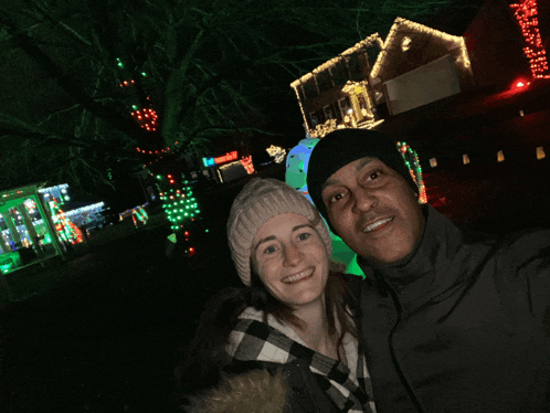 a man and woman are posing for a picture in front of a house with christmas lights on it