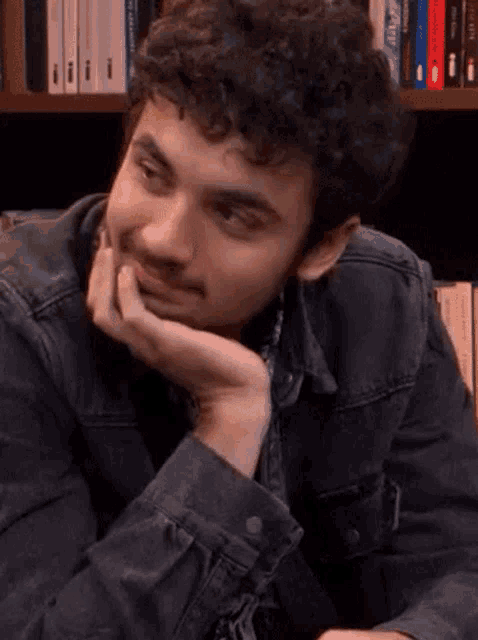 a man with curly hair is sitting in front of a bookshelf with his head resting on his hand .