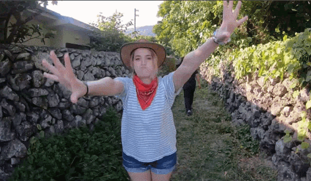 a woman wearing a cowboy hat and a bandana is standing in front of a stone wall with her arms outstretched