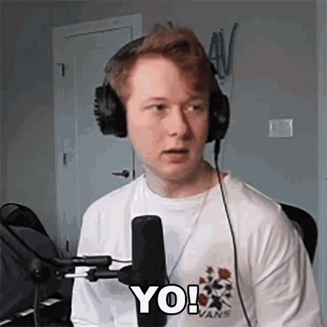 a young man wearing headphones and a white shirt is sitting in front of a microphone and saying yo !