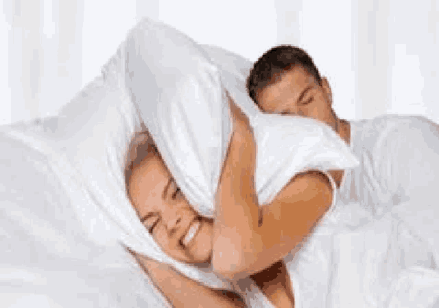 a woman is covering her ears with a pillow while a man sleeps next to her .