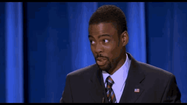 a man in a suit and tie with an american flag on his chest stands in front of a blue curtain