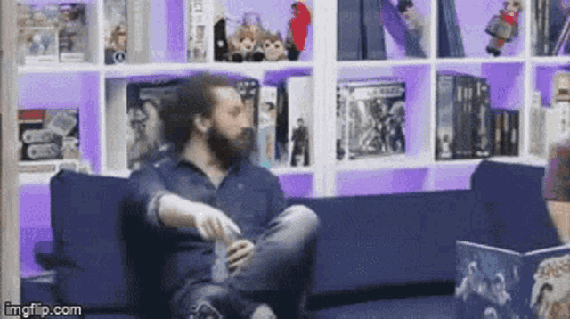 a man with a beard is sitting on a blue couch in front of a bookshelf filled with books .