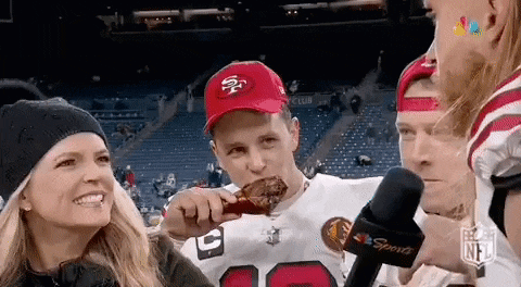 a man in a 49ers hat is eating a chicken wing at a football game while a woman looks on .