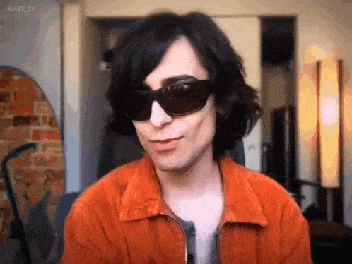 a young man wearing sunglasses and an orange jacket looks at the camera .