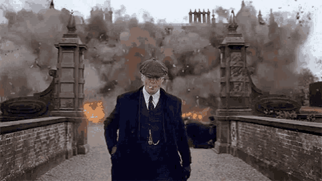 a man in a suit and hat is smoking a cigarette while standing in front of a building that is on fire .