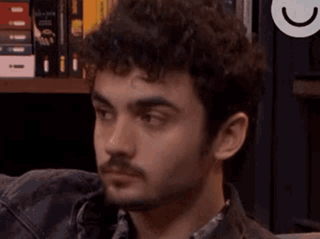 a young man with curly hair and a beard is sitting in front of a bookshelf with his eyes closed .