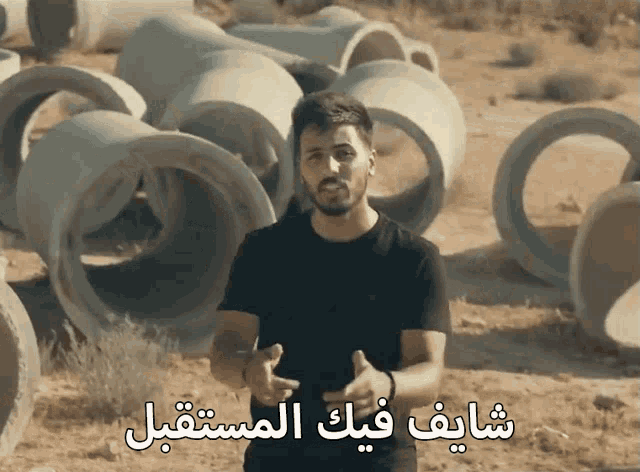 a man in a black shirt stands in front of a pile of concrete pipes with arabic writing below him