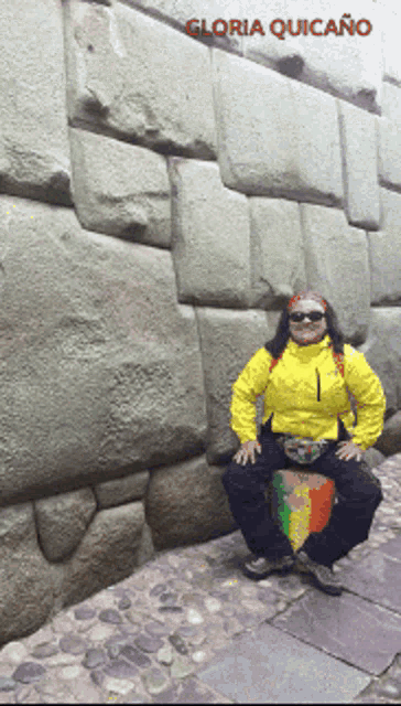 a woman in a yellow jacket is squatting down in front of a stone wall with the name gloria quicaño written on the bottom