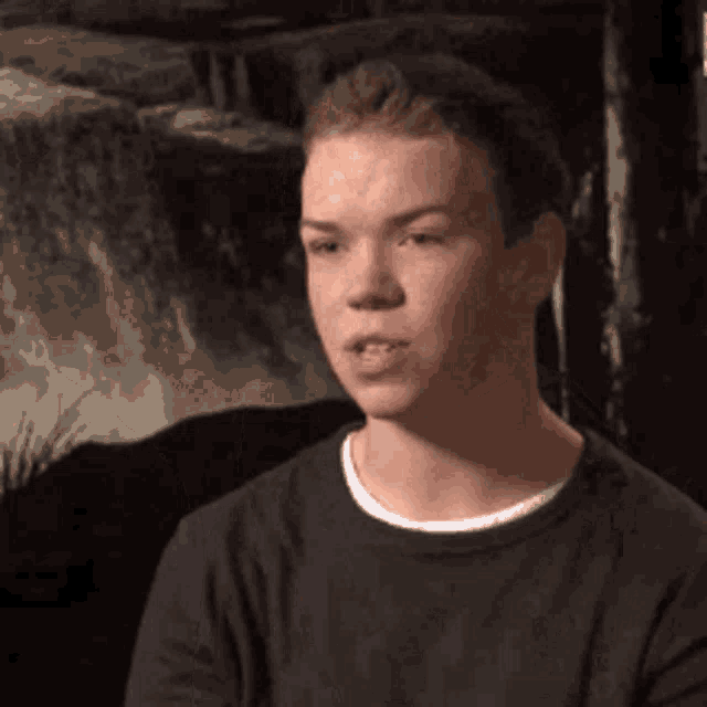 a young man in a black shirt is sitting in front of a waterfall and making a funny face .