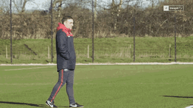 a man in a red and black jacket stands on a soccer field with a swans tv logo in the background