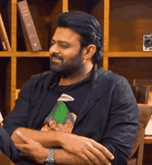 a man with a beard is sitting at a table with his arms crossed and a bookshelf in the background .