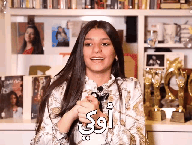 a woman stands in front of a shelf full of books and trophies and says " oui " in arabic