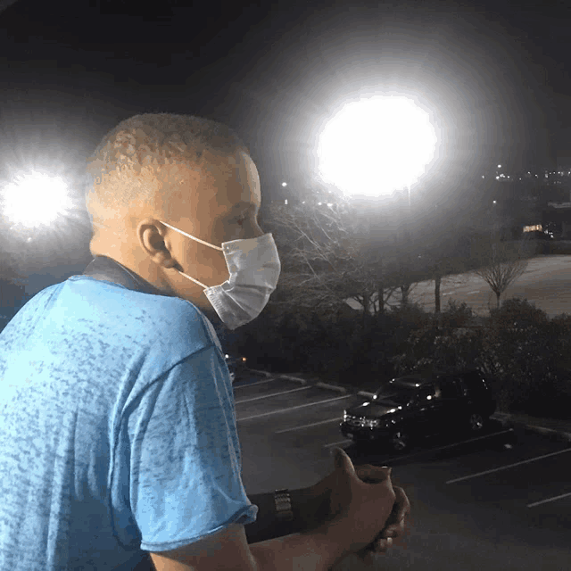 a man wearing a blue shirt and a face mask stands in a parking lot at night