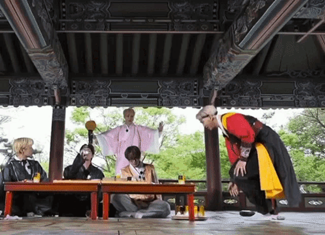 a group of people are sitting at tables under a gazebo while a man in a red and yellow outfit stands in front of them