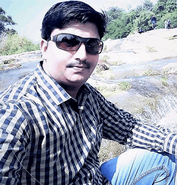 a man wearing sunglasses and a plaid shirt sits on a rock near a river