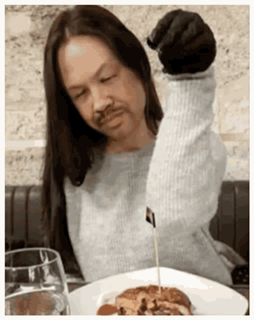 a woman with long hair and a fake mustache is sitting at a table with a plate of food and a glass of water