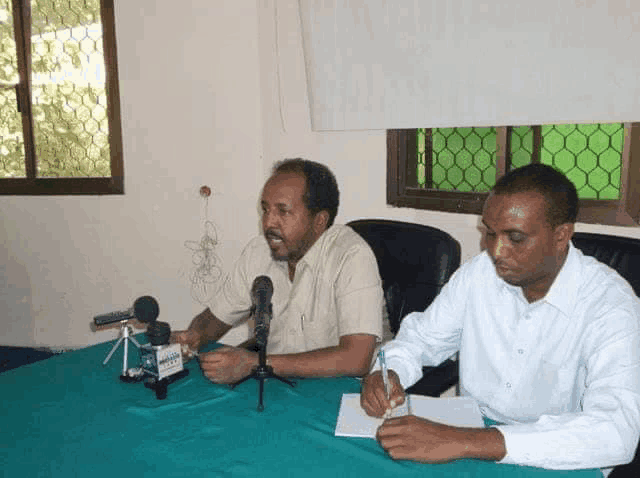 two men are sitting at a table with microphones and one of them is writing on a piece of paper with a pen