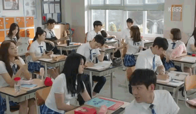 a group of students are sitting at desks in a classroom with kbs2 written on the wall behind them .