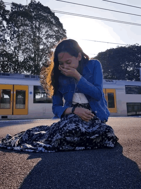 a woman covering her mouth while sitting on the ground in front of a train with a yellow door that says a