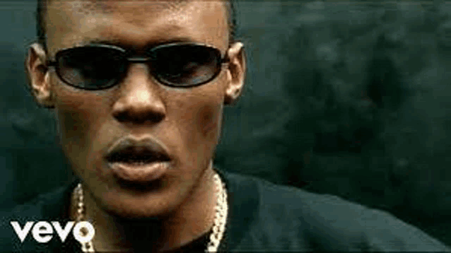 a man wearing sunglasses and a gold chain is standing in front of a black wall .