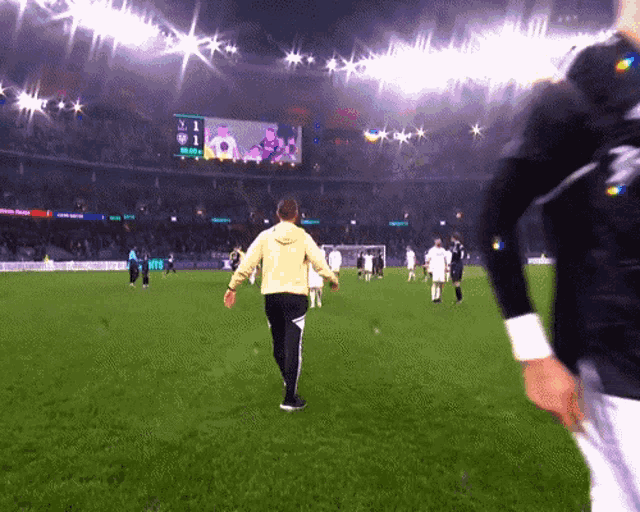 a man in a yellow hoodie walks on a soccer field with a scoreboard in the background that shows the score of 0-1