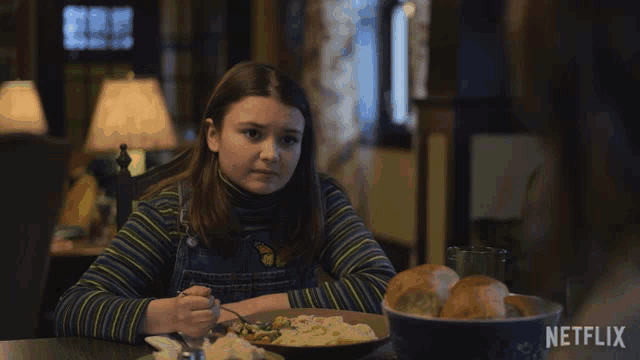 a girl sits at a table with a bowl of bread and the words " never mind " on the screen