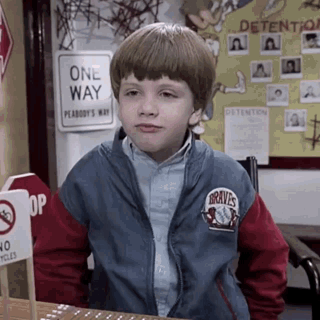 a young boy wearing a braves jacket is sitting at a desk in front of a one way sign .