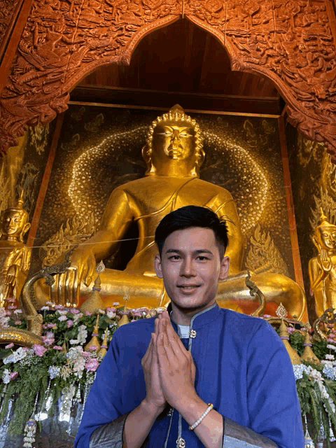 a man in a blue shirt is praying in front of a large gold buddha statue